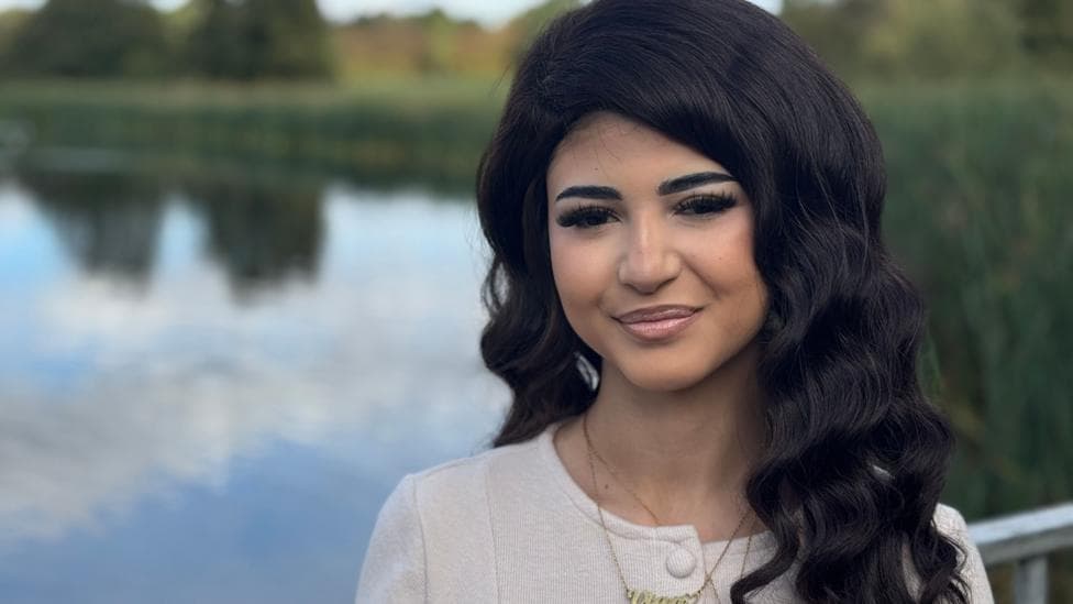 Vian stands next to a lake looking into camera. She has long dark hair and is wearing a light coloured top and necklace.