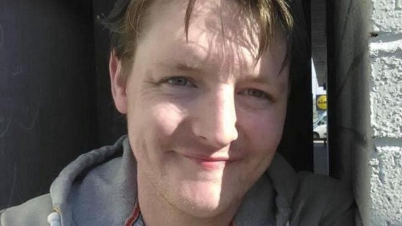 Jamie Forbes leaning against a white wall, smiling at the camera. He is wearing a grey hooded jumper.