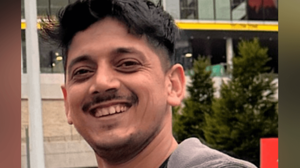 A man with short black hair and a short stubbly moustache and beard wears a grey zipped hoodie and black T-shirt while smiling for a photo.