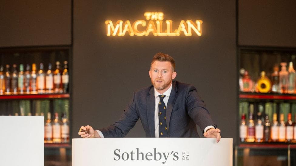 A man stands at an auction block in a suit in front of shelves of bottles. The auction block says Sotheby's