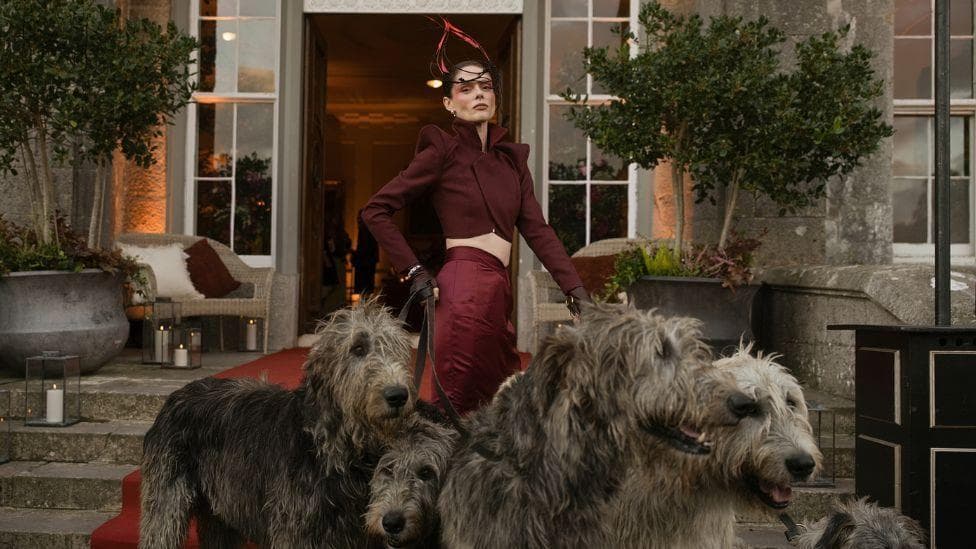 A woman wearing an elaborate red outfit, with a headpiece, holding multiple large Irish wolfhounds on leads. She is stood on the steps of a grand looking building with large potted plants and full length windows