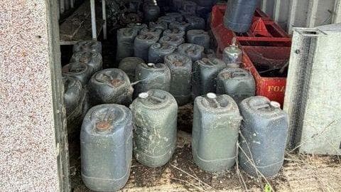 A number of dark green containers inside what appears to be an open building. There are two red crates on one side. The building has grey walls and there appears to be an earth floor.