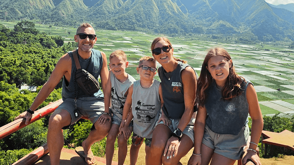 Family of five sitting on a wooden fenced ledge with rice padis in the background and green hills behind . Tom is 38, white, short fair hair. Morgan and George have short fair hair. Dani has long fair hair in a ponytail and Olivia has loose long dark blonde hair. All five wear shorts and sleeveless tshirts in varying shades of grey. Dani and Tom wear sunglasses. They all smile at the camera.
