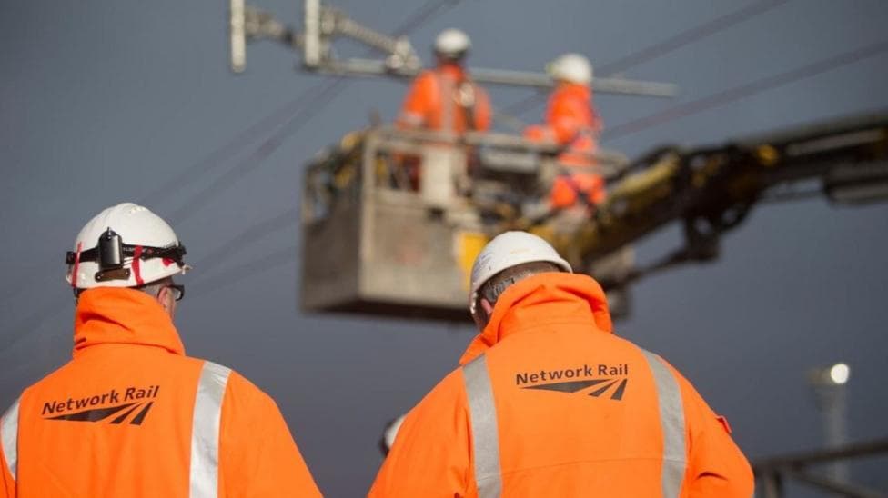 A photo showing the backs of two men in orange hi-viz jackets which say Network Rail on the back of them. They also wear white hard hats. In the background, two men are in a cherry picker.
