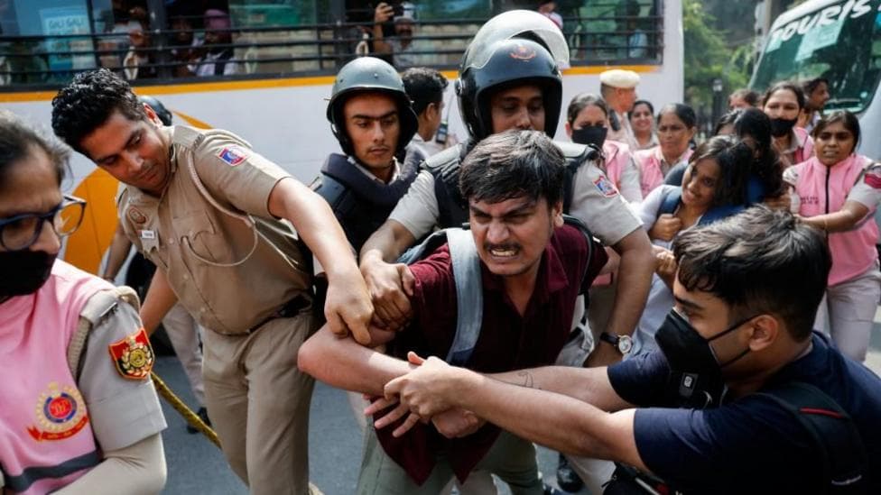 A man is being held by men in official uniforms as another man grapples with them. More people are stood behind in pink uniforms holding other protesters.