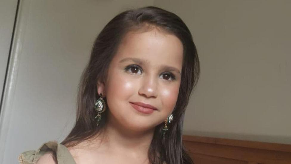 A 10-year-old with dark hair and eyhes looks forward, smiling with her mouth closed. She wears make-up and earrings.