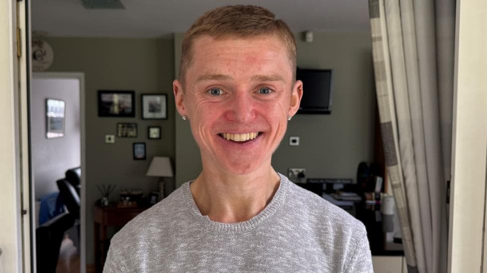A man smiling at the camera. He is wearing a grey jumper