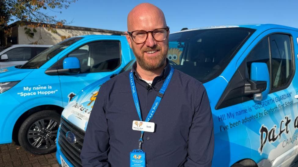 Scott Jobson, a man with a shaved head and beard who wears glasses, standing in front of the bright blue vans used by the Daft as a Brush charity. He is wearing a similarly coloured lanyard.