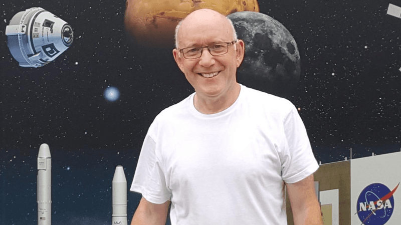 A bald man with glasses wears a white t-shirt in front of a poster about space.