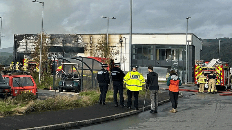 A burnt factory - there are police officers in front and a fire engine. There is a car park to the left.
