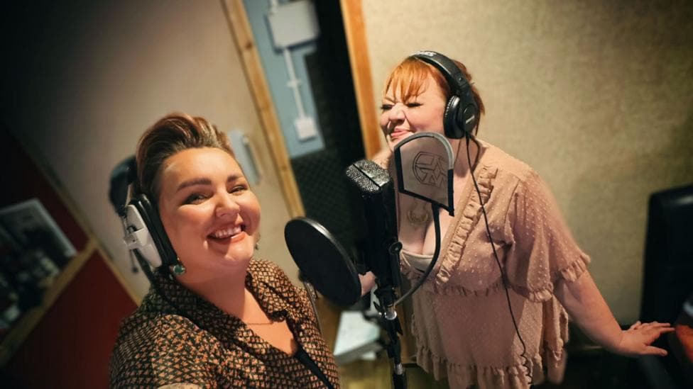 Bronwen Lewis (left) and Jada Star (right) recording the Welsh version of Jolene in Southampton. Lewis who has short hair is wearing headphones whilst smiling and looking at the camera. Star whose red hair is tied up is smiling with her eyes closed. She is also hearing headphones. The pair are stood with a microphone between them.