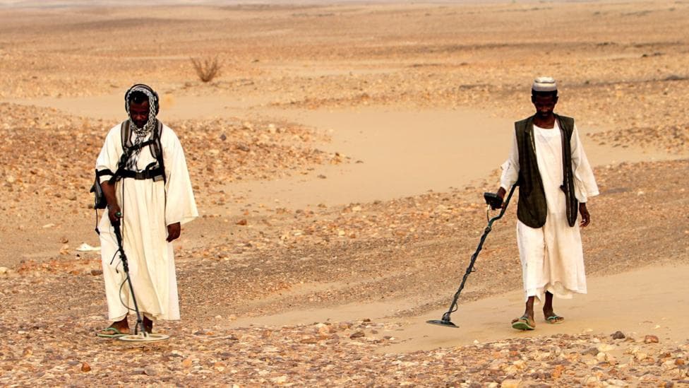 Sudanese men wearing traditional clothing search for gold with metal detectors in the desert.