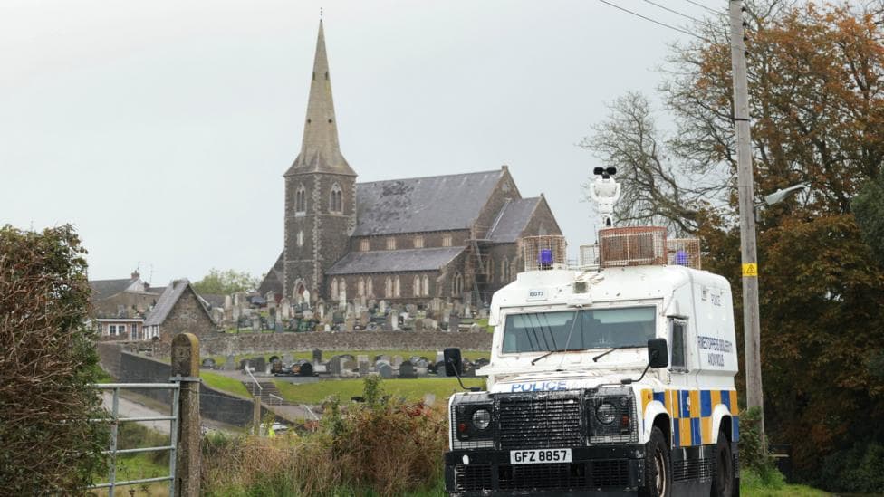 A PSNI land rover close to Drumcree Church in Portadown. It's white with metal guards at its front and over its sirens. There is a blue and yellow checkered pattern around it just above the wheels. It is in front of a grey stone church. The sky is cloudy. The PSNI land rover is parked at a side of a road, Drumcree church is seen in the background where a graveyard is also visible.