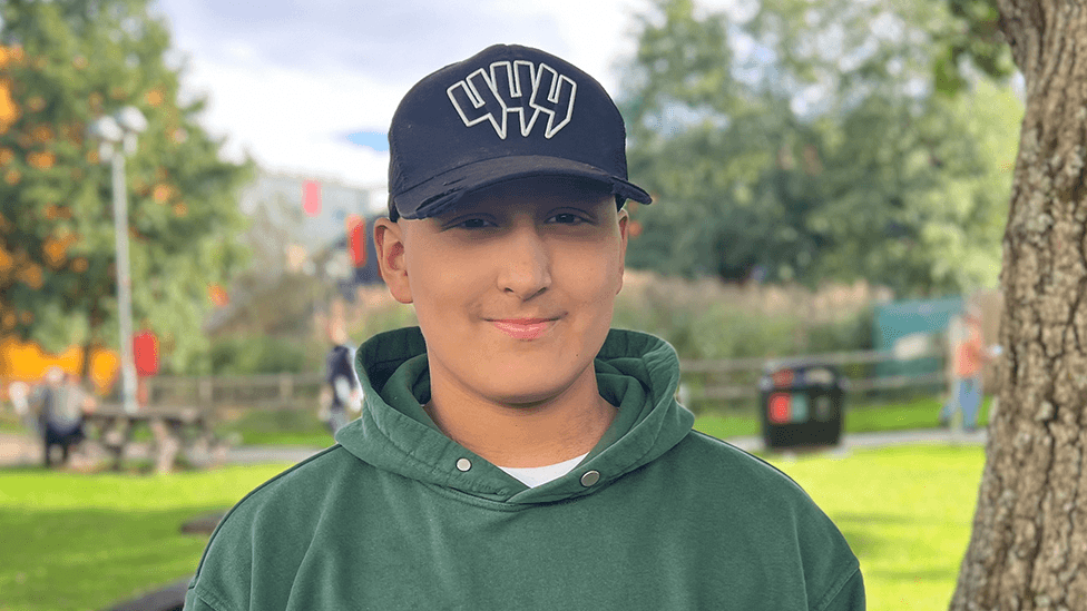 Ethan looks at the camera and is wearing a dark green hoodie and a black baseball cap with a white logo. He is standing outdoors beside a tree. Grass, more trees, picnic benches and several people are visible out of focus in the background.