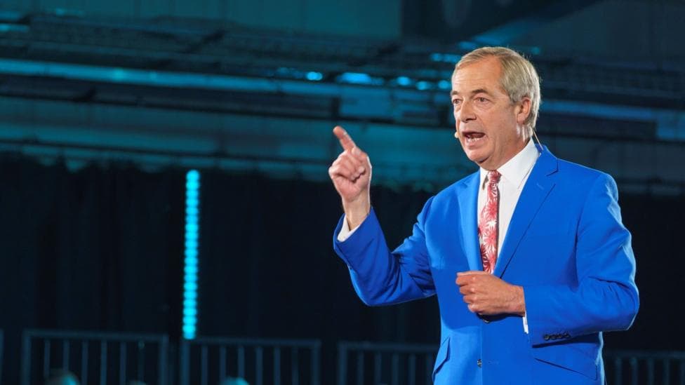 Nigel Farage wears a blue suit, white shirt and red tie as he speaks on a stage, wearing a microphone.