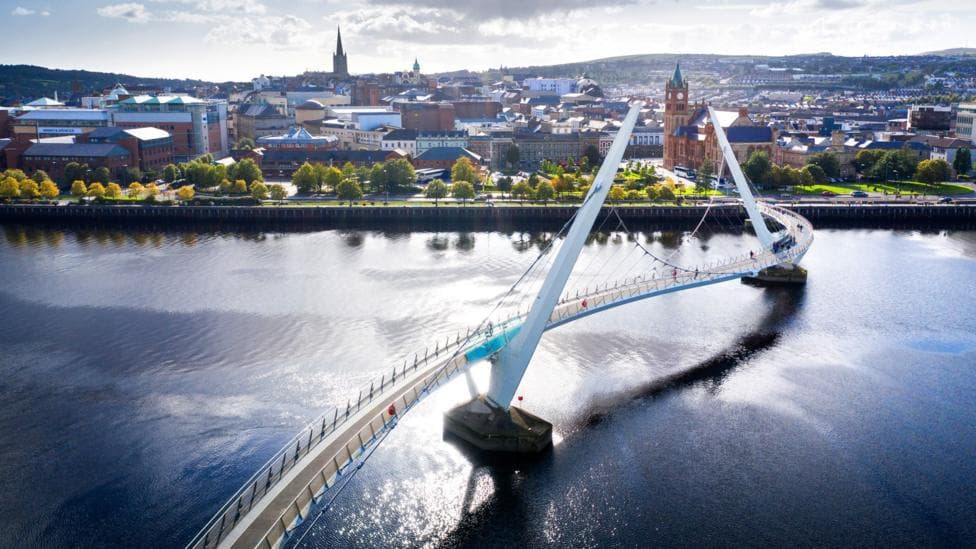 An aerial view of the peace bridge in Derry, as it spans the river Foyle. In the background is the city, with the Guildhall and other buildings visible.