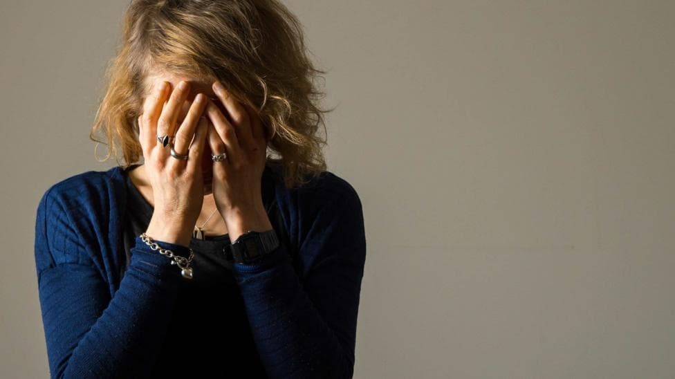 A person is seated against a plain, light-coloured wall with both hands covering most of the face. The person is wearing a dark blue long-sleeved top, a wristwatch, several rings and a bracelet, with medium-length hair falling forward, while soft lighting from the left side of the image creates shadows across the figure and the otherwise empty background.