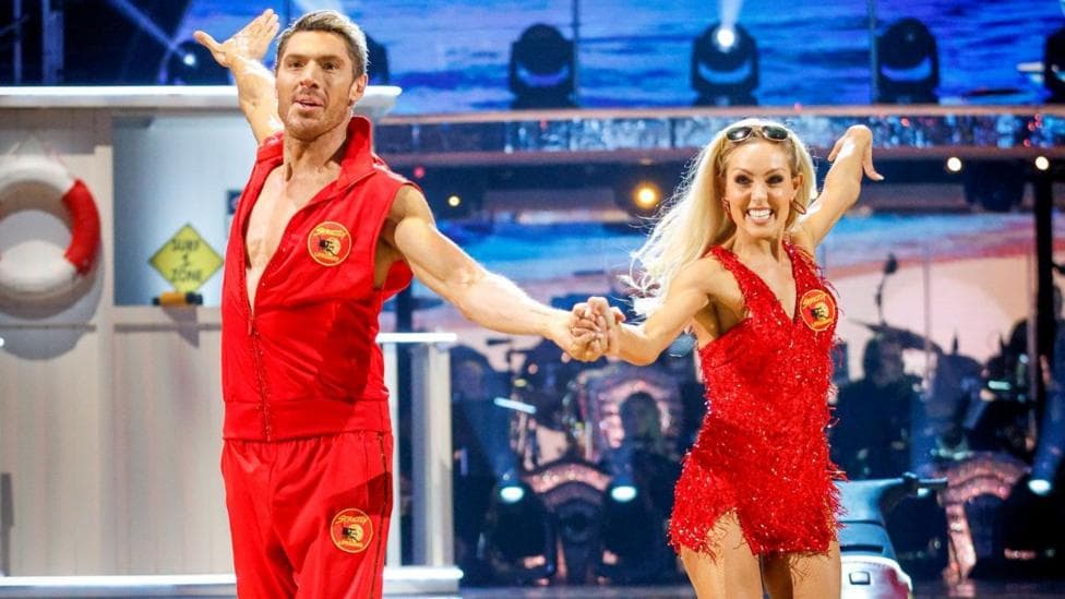 Amy Dowden and Chris Appleton during their appearance on the live show on Saturday 3 October for BBC1's Strictly Come Dancing. Dowden and Appleton wear red Baywatch outfits and are dancing hand in hand on the Stricty dancefloor. Amy has long blonde hair and a red sparkling short dress. Appleton wears a red waistcoat and red joggers and has blonde short hair. They both smile.