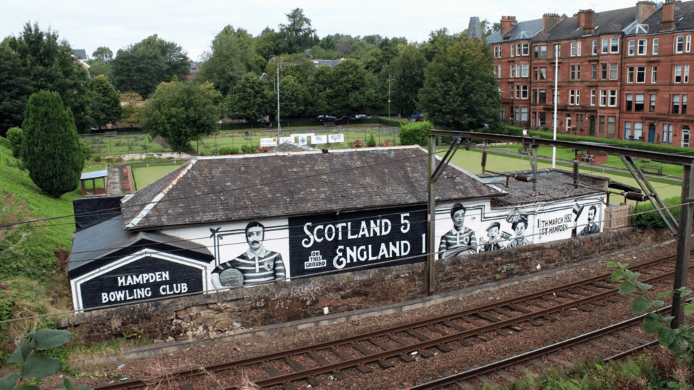 A bowling green ground from across train tracks behind it. A mural on the back wall says Scotland 5 England 1 with images of players from the 1880s alongside it. Flats can be seen in the background behind the green.