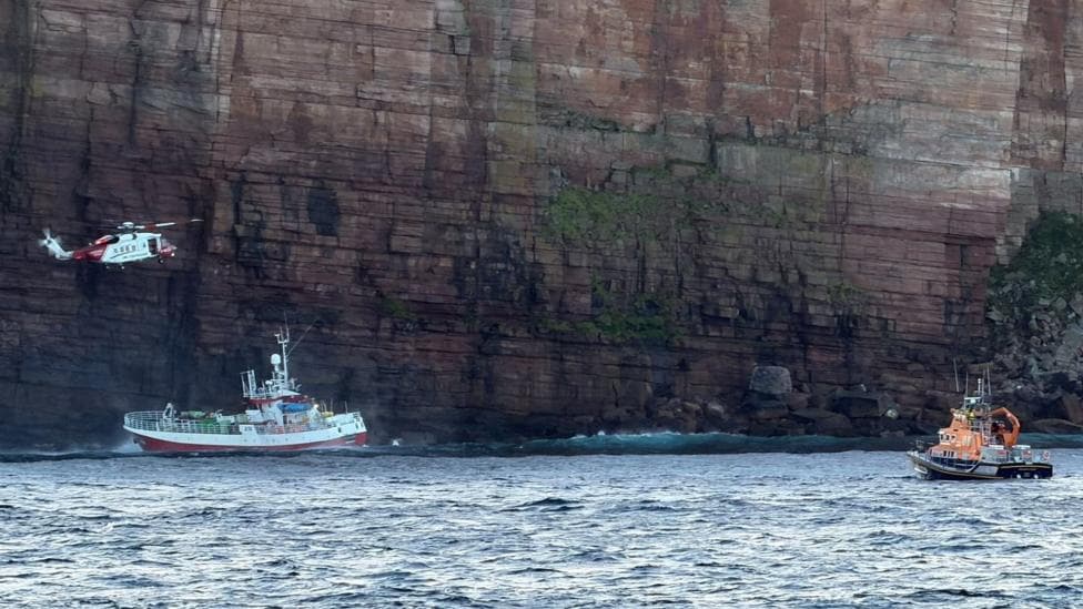 A rescue helicopter hovers beside a fishing boat near towering red sandstone cliffs, while a lifeboat waits nearby on the sea.