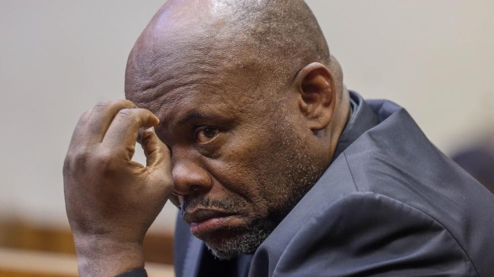 A close-up shot of Shadrack Sibiya gesturing with his left hand. He has a shaved head and is wearing a dark suit