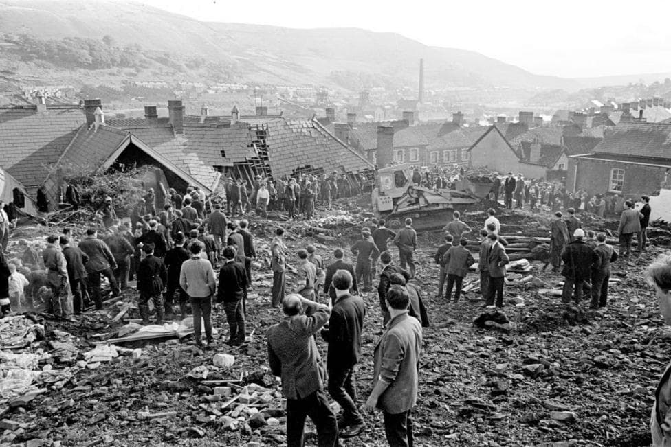 A black and white photo showing rescue workers standing and surveying the damage to the ruined junior school in Aberfan. Coal slurry and debris covers the floor.