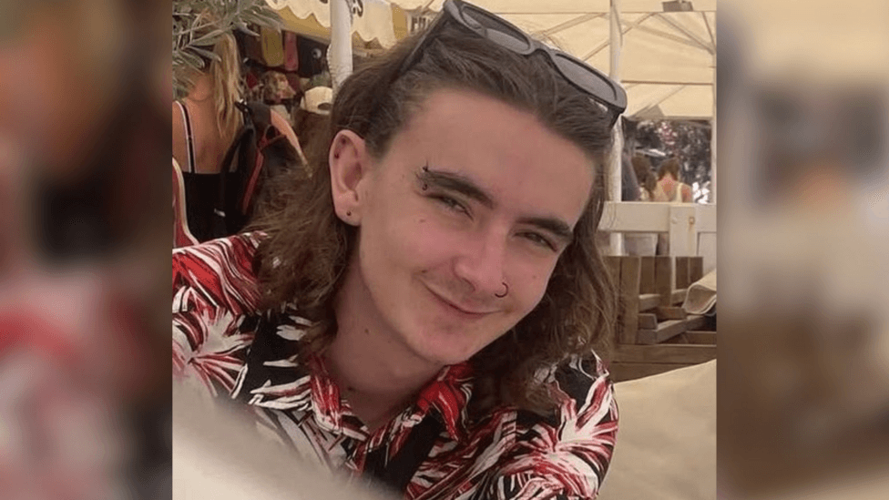 Jamie Collins wearing a black, red and white patterned shirt and sunglasses on his head. He has shoulder length brown hair and piercings in his ears, eyebrow and nose. He appears to be sitting underneath a white marquee on a sunny day.