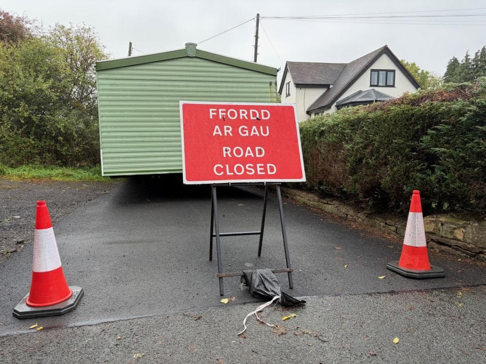 A green static caravan blocking the road. There is a bilingual sign saying "Fford are Gau/Road closed" and two traffic cones.