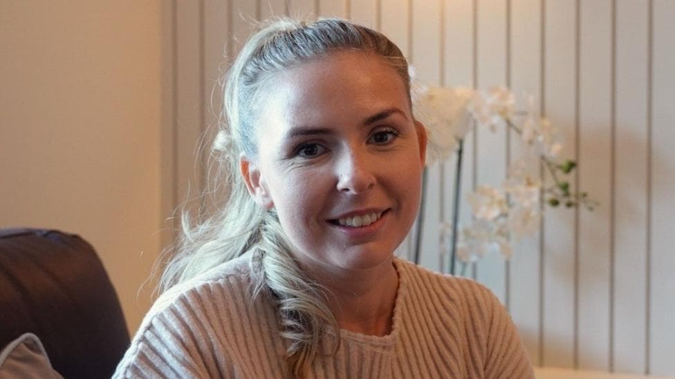Kelly O'Shea sat in her living room, against a backdrop of a cream wall, with an orchid on the shelf behind her. She is wearing a cream ribbed top and has blonde hair tied back from her face.