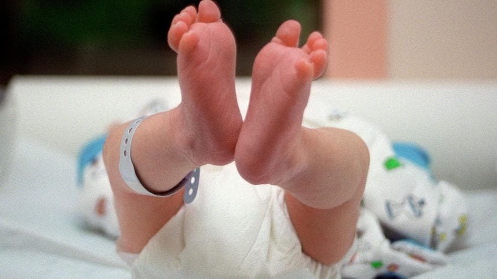 Image showing a newborn baby lying down with the soles of their feet in the foreground. The baby is wearing a nappy with a tag attached to one leg.