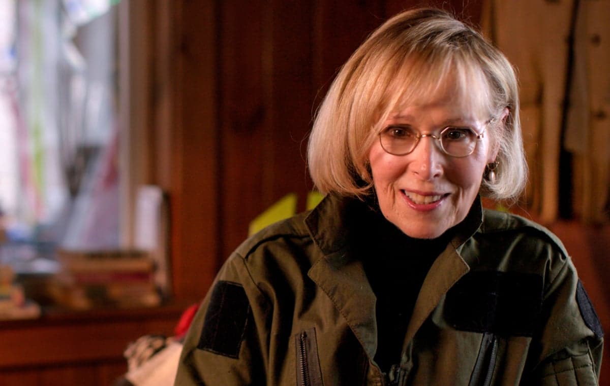 A woman with a blond bob haircut, gold-rimmed glasses and a brown jacket