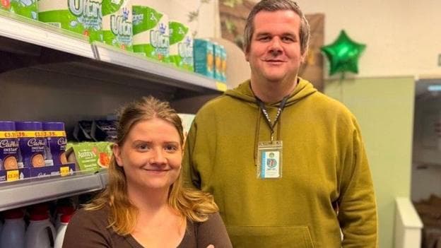 A tall man and short women both looking at the camera smiling. They are stood in front of a shelf of various food and drink items.