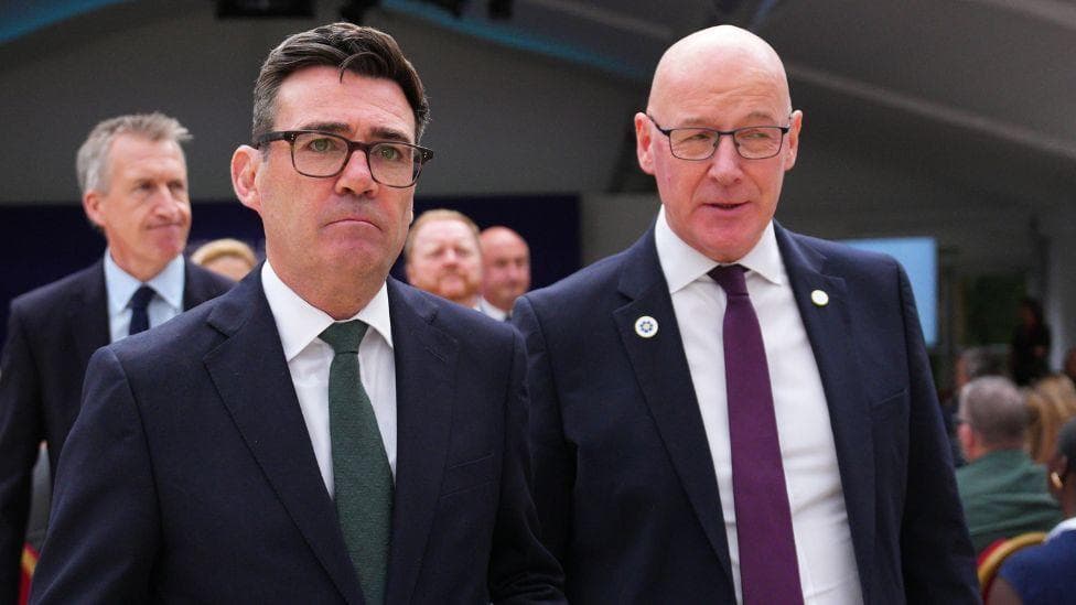 Andy Burnham, who has dark hair and glasses, and John Swinney, who is bald with glasses, walk side by side. They are both wearing business suits