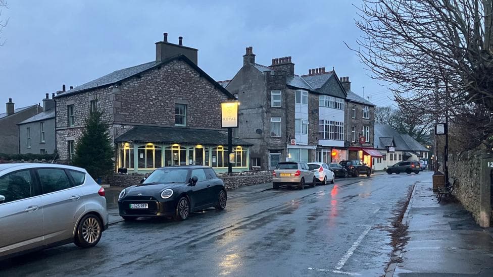 A photo of Silverdale, in Lancashire. The picture shows a street with homes and businesses, and, to the left, the outside of a pub. It is a rainy evening, and the light is fading - there are cars parked along the road.