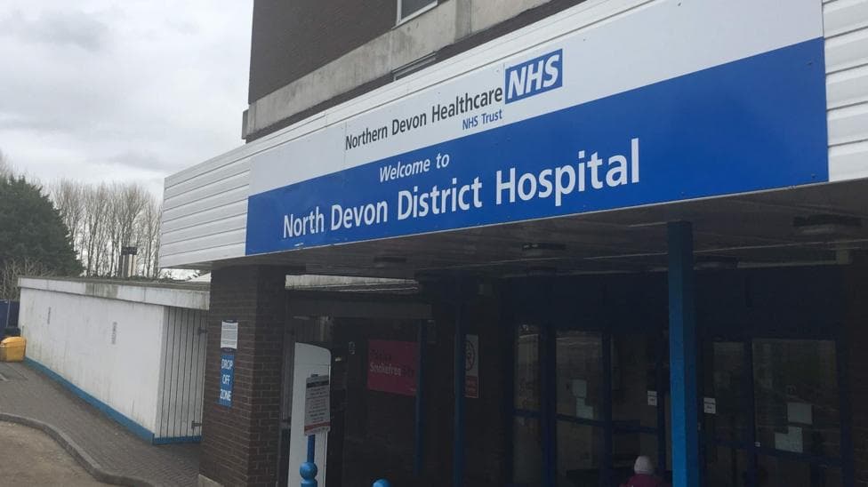 Outside one of the entrances to the North Devon District Hospital site. A blue and white sign with the hospital's name on it are above the main doors.