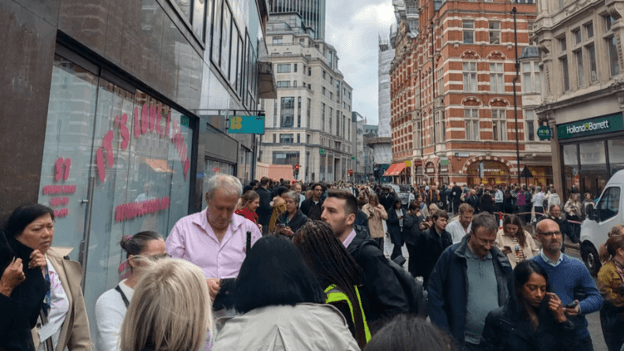 Crowds of people stand on a pavement and spill into the road on a street in the City of London, surrounded by office buildings and shops.