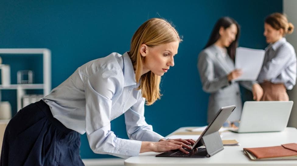 A female office worker wearing a light blue top and dark blue skirt leans over a laptop computer. Two other workers are in the background, slightly out of focus
