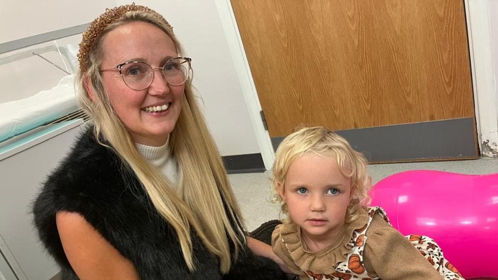 Jo is wearing round glasses, a cream top and a black faux-fur vest. She sits on the floor beside her daughter, Phoebe who has light, curly hair. Phoebe is wearing a patterned pinafore dress over a brown long-sleeved top and holding a red toy block. Bright yellow and pink therapy balls surround the pair in a clinical room with a wooden door and storage unit visible in the background.