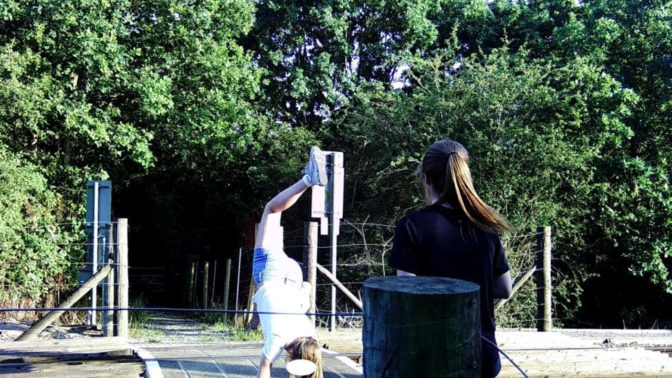 A girl in a white T-shirt and denim shorts does a handstand on the wooden deck of a level crossing as another girl with a ponytail, wearing a black T-shirt, watches with her back to the camera.