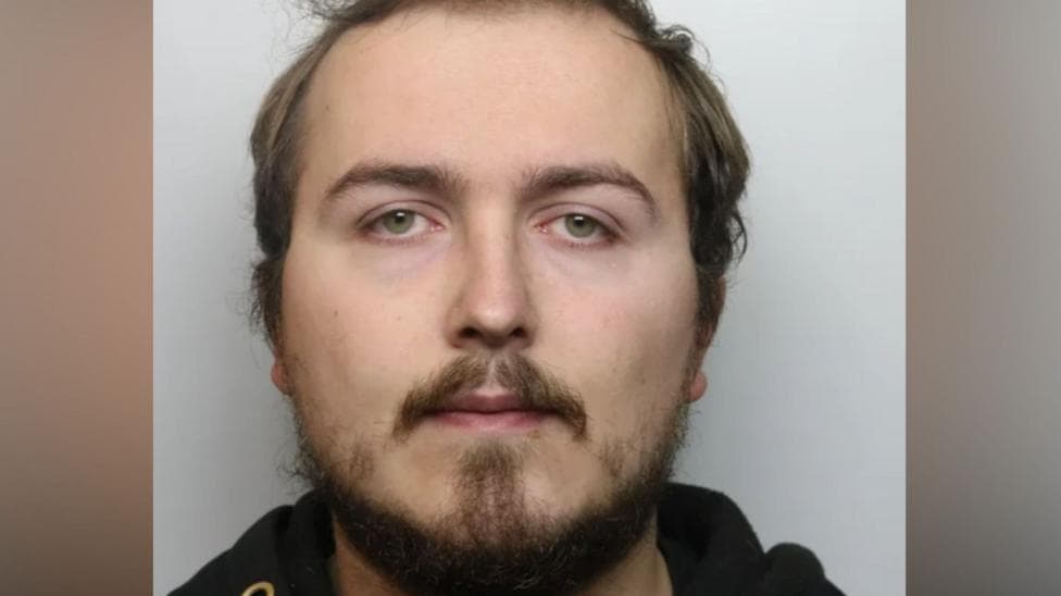 A police mugshot of a man with dark brown hair and beard. He is positioned against a light coloured background.