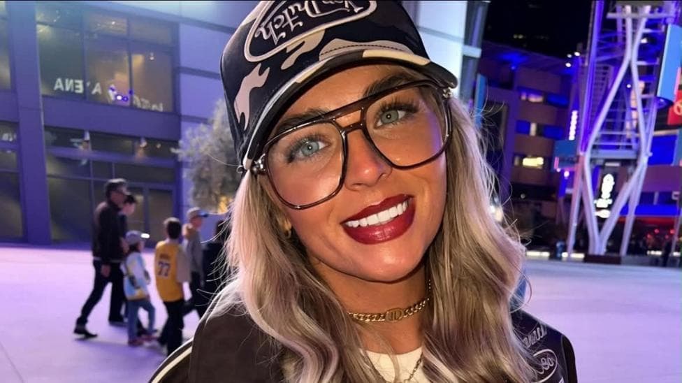 Denby McLaren - a smiling woman with large glasses and a baseball cap, standing outside a sports arena.