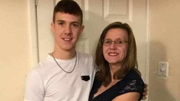 A mum and son are stood in a room looking at the camera in a white room. The man is wearing a white t-shirt and the woman a black dress.