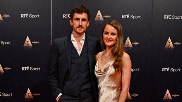 Ciara Mageean with her partner Thomas Moran. They are at a red-carpet event, and she is wearing a gold gown with her hair swept to one side. Thomas is wearing a three-piece navy suit. He has dark hair and a beard.