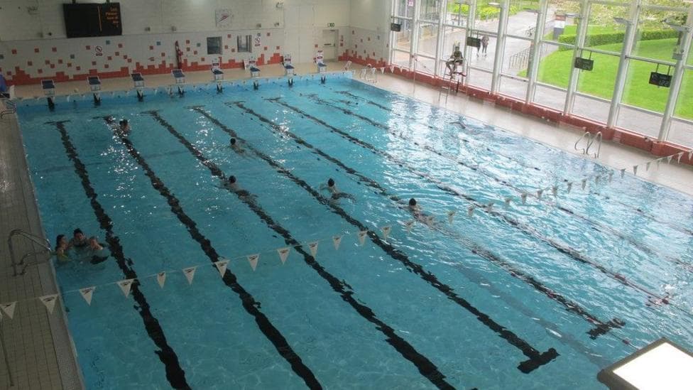 Large indoor swimming pool with lanes marked out in black on the pool floor. Bunting stretches above the pool at one end. People are in the water. A lifeguard sits on a high stool at the side of the pool near large windows which look out to the campus with green lawns and hedging.