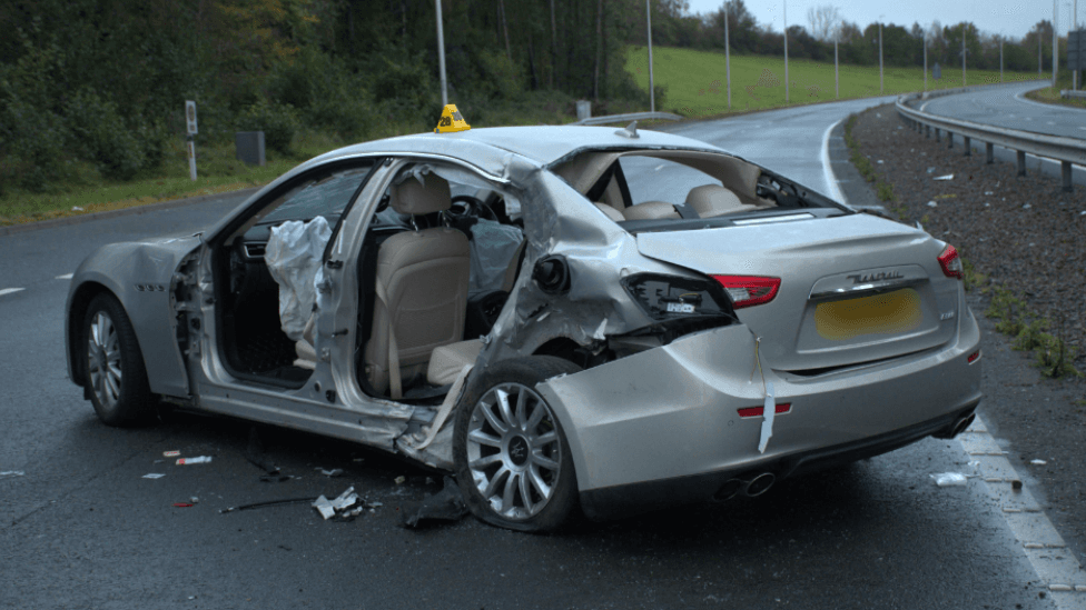 A behind view of a grey Maserati car in the middle of a lane on a dual carriageway. The car is visibly damaged with the air bags blown up and the doors missing. There is debris around the car on the road.