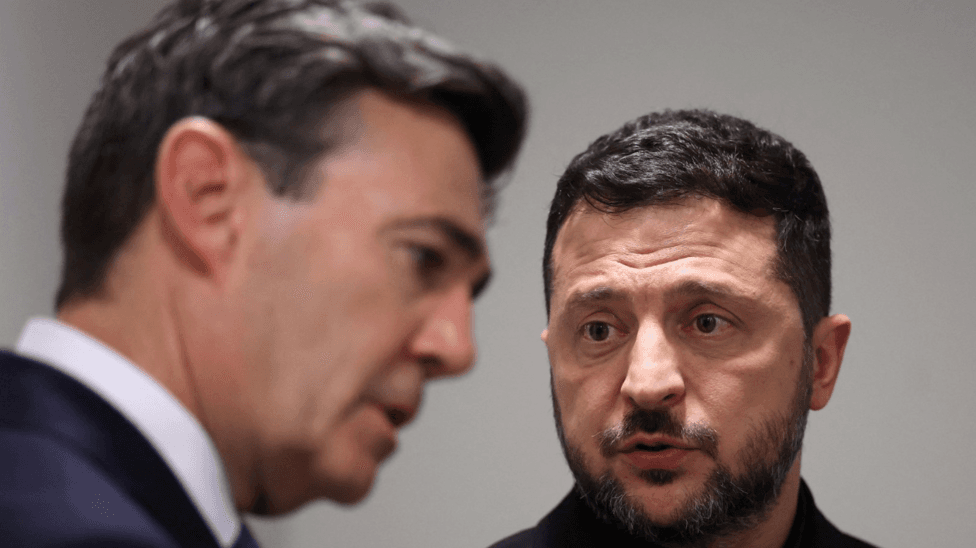 A head and shoulders shot of Zelensky on the right, who has a close-cropped beard and is wearing a black top. Front right is an out of focus shot of Burnham, wearing a navy suit and white shirt with a tie, and listening intently.