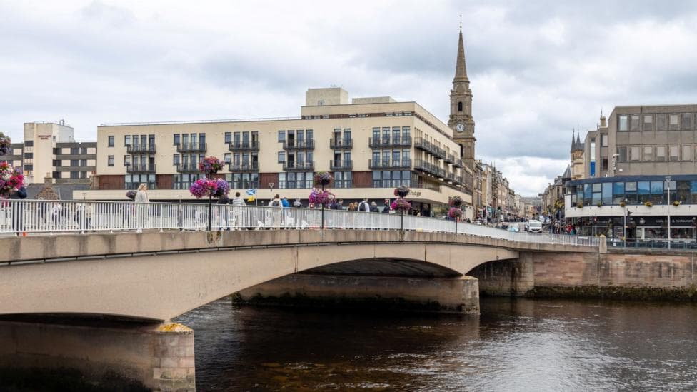 A view along Ness Bridge over the River Ness towards the area of the junction. There are rows of buildings and a steeple with a clock. Cars are just visible on Bridge Street.