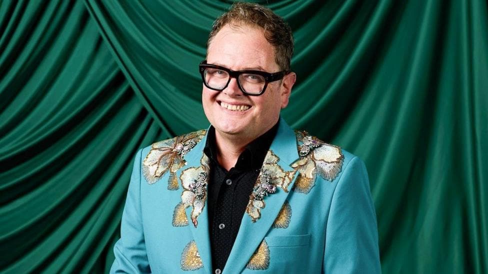 Comedian Alan Carr with his trademark black-rimmed glasses in a blue suit jacket with flowers on it and a dark shirt underneath. He is standing in front of a green curtain.