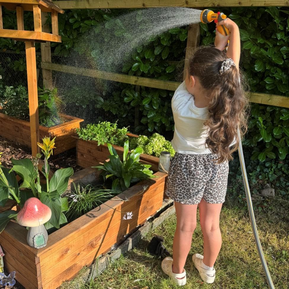 Child standing in a garden in the foreground, holding a hose spray nozzle above their head and watering raised wooden planters. Water arcs from the nozzle across the planters, which contain various flowers and leafy plants. A wooden bird-table-style structure stands beside the planters, and a mushroom-shaped garden ornament is visible at the corner of one planter. A wooden fence and dense hedging form the background.
