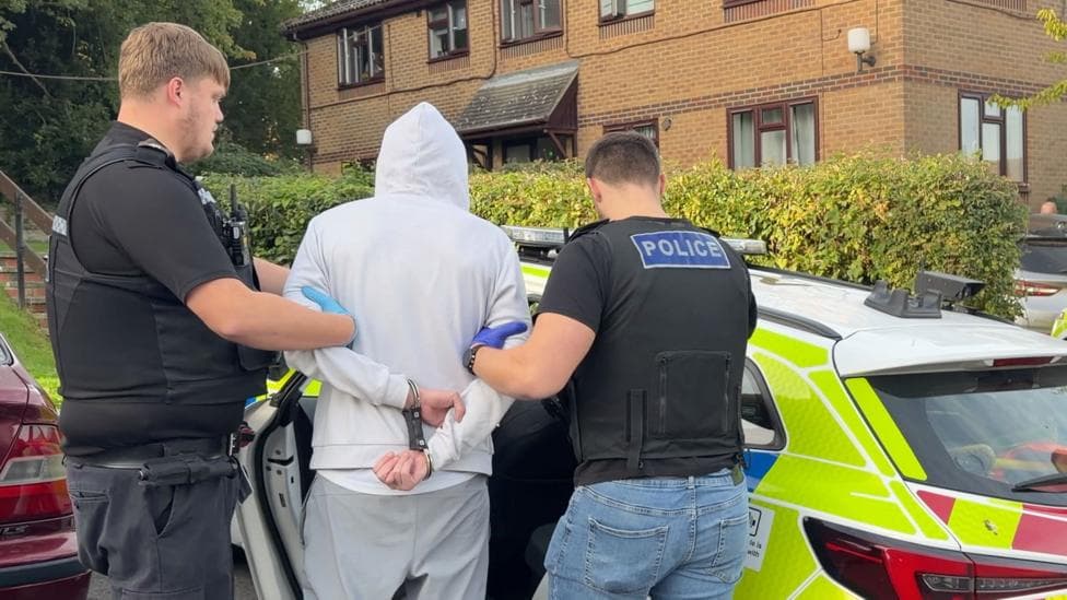 Police officers in plain clothes with police stab vests on top lead a handcuffed suspect wearing a hoodie into a police car, which is parked outside a low-rise, brick-built block of flats.